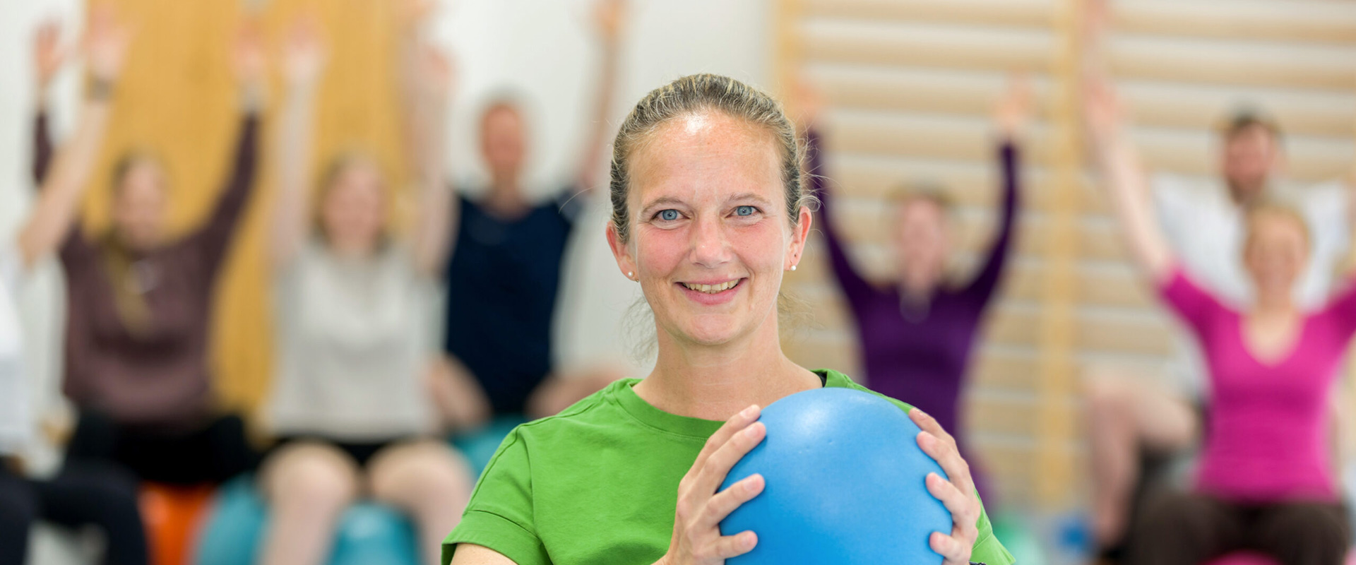 Eine Frau lächelt freundlich und hält einen blauen Ball in die Luft. Im Hintergrund sieht man Menschen, die die Arme heben.