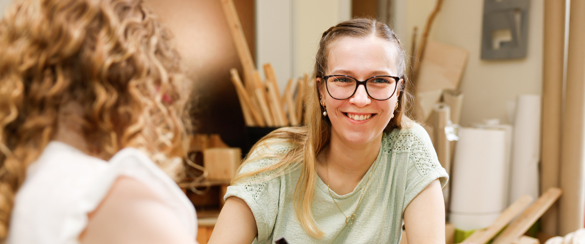 Eine Frau mit Brille lächelt freundlich. Sie befindet sich in einem Werkraum. Ein Kind, welches nur zur hälfte und von hinten zu sehen ist, hat einen Hammer in der Hand.